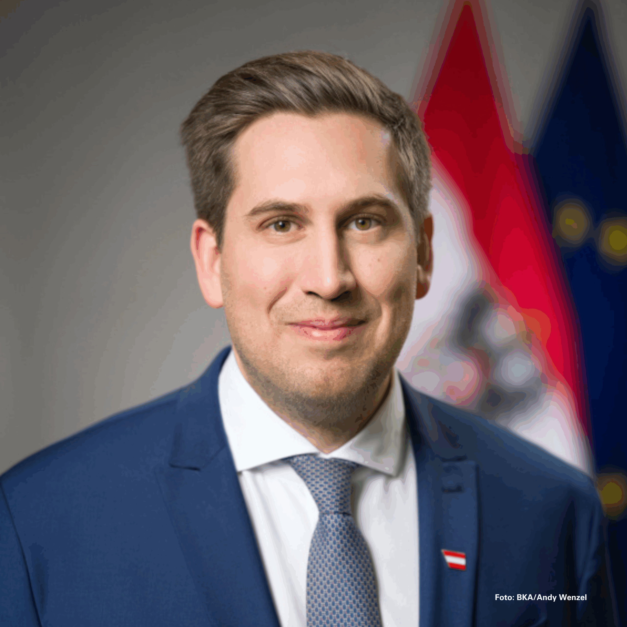 Er hat braune Haare, trägt einen blauen Anzug mit weißem Hemd und hellblauer Krawatte